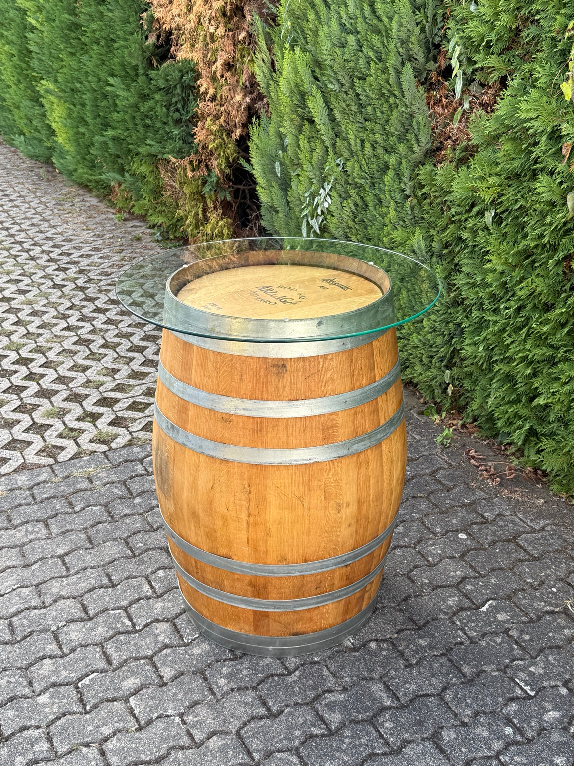 Weinfass-Stehtisch aus geölter Eiche mit runder Glasplatte auf gepflastertem Boden vor einer Hecke
